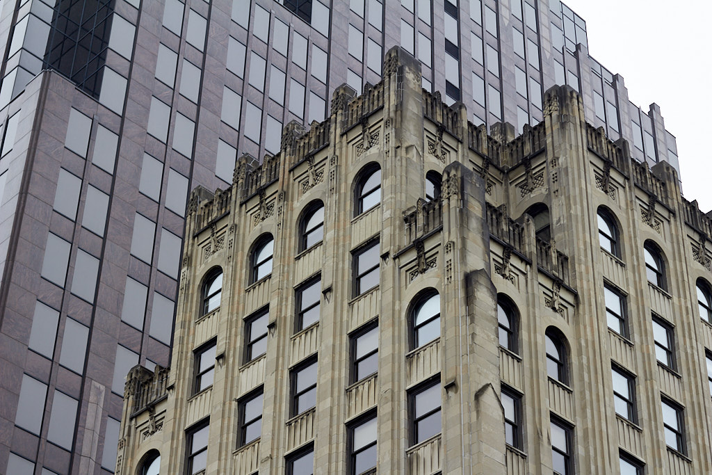 Photograph with muted colors of the top of an office building with a limestone façade and art deco ornamentation stained dark by acid rain in the foreground and a larger, darker, more modern office building extending skyward in the background.